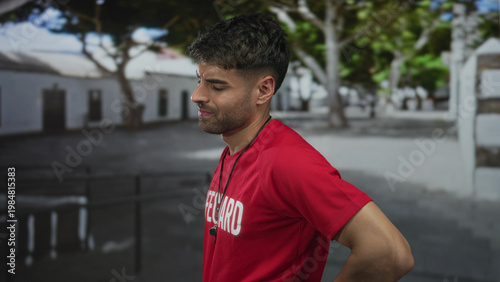 Fotografie Young man in red lifeguard shirt with whistle holds lower back and looks sideways on street; backache discomfort