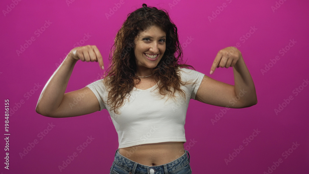 Obraz premium Woman pointing both index fingers down at her exposed navel in a magenta studio, wearing a white cropped shirt and jeans; playful confidence.