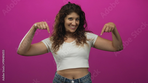 Woman pointing both index fingers down at her exposed navel in a magenta studio, wearing a white cropped shirt and jeans; playful confidence.