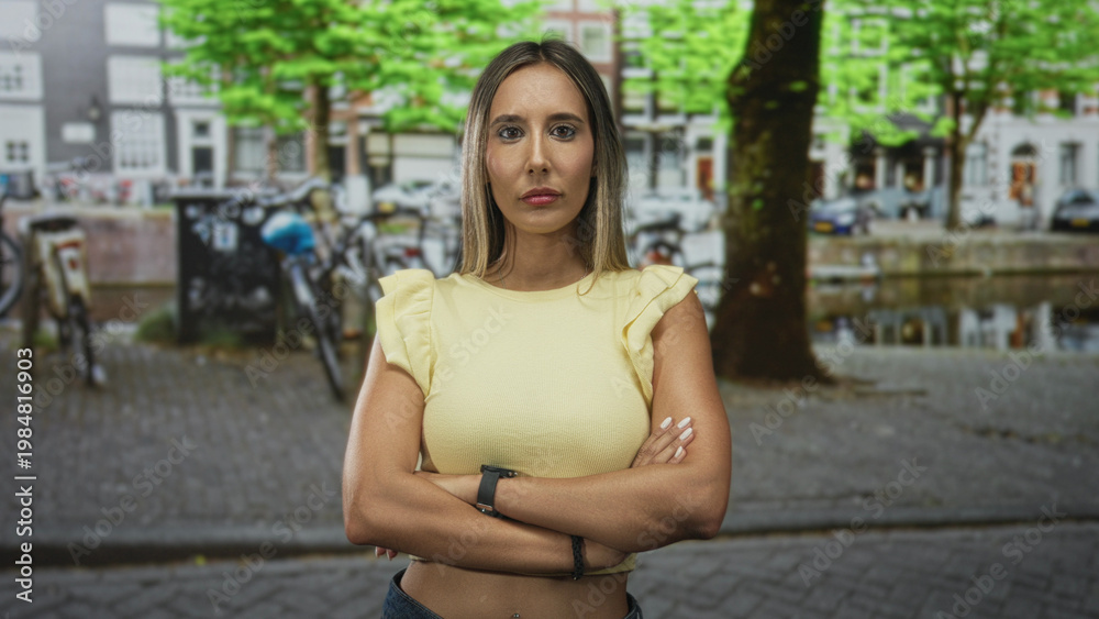 Fototapeta premium Young hispanic blonde woman with arms crossed wearing a yellow shirt on a cobbled street by a canal with parked bicycles and a large tree; determination independence.