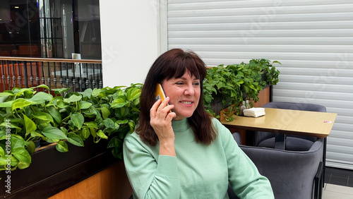 Middle-aged woman sitting in a coffee shop talking on her mobile phone and smiling. Concept of communication, friendly conversation, and relaxed lifestyle. Social connection, leisure time, and modern