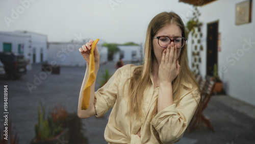 Quadro su tela Woman holding yellow sock on a street with outdoor chairs while sniffing odor with a grimace; disgust