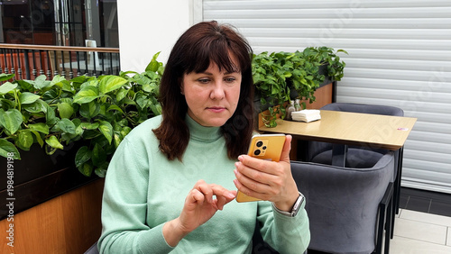 Middle-aged woman sitting in a coffee shop using her smartphone. Concept of communication, online shopping, and digital entertainment. Modern lifestyle, mobile technology, and leisure time in a cafe.