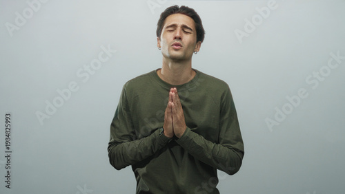 Tablou pe pânză Man with hands pressed together in prayer gesture, closed eyes and pleading lips, wearing olive long sleeve shirt and wristwatch against a neutral grey studio backdrop; plea hope