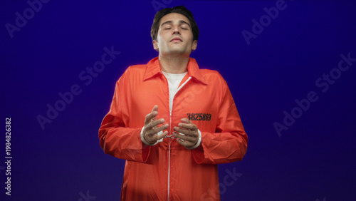 Fotografia Man in orange prisoner jumpsuit showing inmate number with clasped hands and closed eyes in studio; remorse reflection solitude