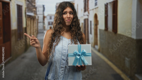 Canvas-taulu Woman holding gift box with blue ribbon in an old street, sleeveless top and curly hair, offers it with hesitant expression; reluctant offering uncertainty