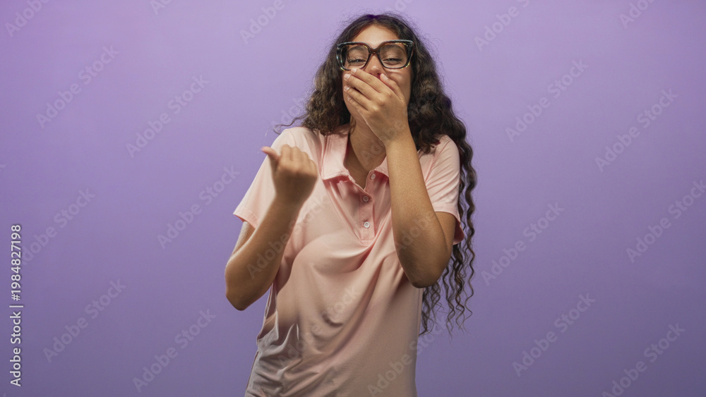 Fototapeta premium Hispanic teenage woman in pink polo covers mouth with hand while laughing and pumping fist in studio; playful amusement.