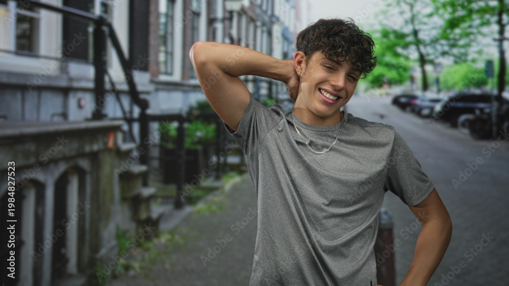 Fototapeta premium Teenage boy with hand behind neck smiling in gray t shirt on a street by a building; playful youth moment.