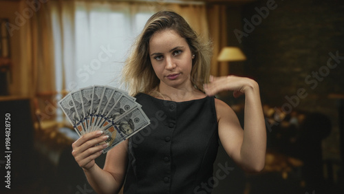 Obraz na płótnie Woman holding a fan of hundred dollar bills and flipping her hand in a studio living room, seated near a lamp and curtains; confidence wealth