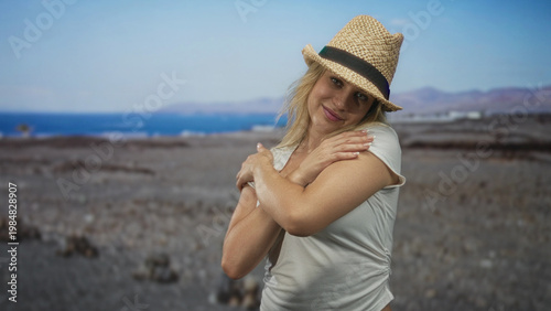 Young woman with straw hat and white tee, arms crossed over chest hugging herself on coastal shore; serenity.