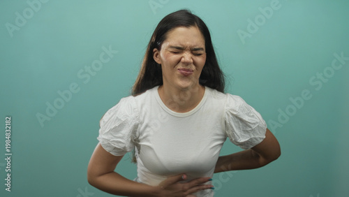 Foto Young hispanic woman holds lower back and presses stomach while grimacing in teal studio; discomfort