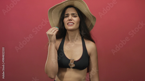 Woman wearing wide brim straw hat, black bikini top and turquoise nails pinches nose and scowls in studio; disgust.