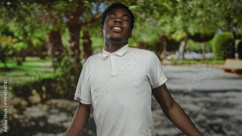 Man in white polo shirt stretches arms in green park with eyes closed and head tilted back; relaxation acceptance openness growth.