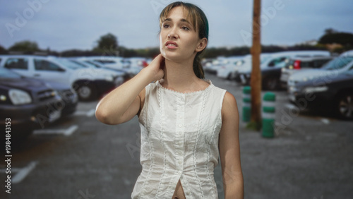 фотографія Young woman in white sleeveless top touching neck and exposed midriff in street among parked cars; frustration waiting