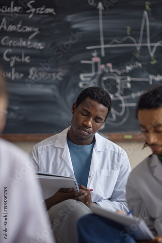 Vertical shot of young Black male medical student taking notes during anatomy lesson, studying cardiovascular system. Useful for healthcare education, training, university marketing