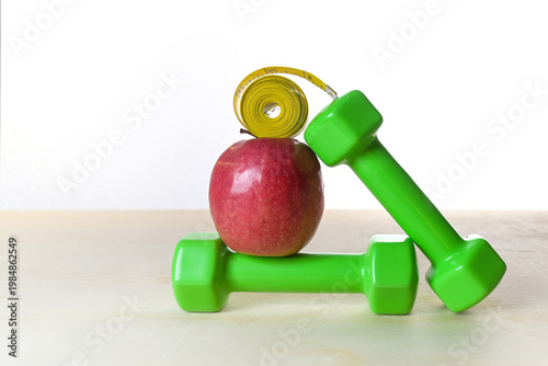 Dumbbells, apple and a tape measure stacked in balance on light wood against a white background, healthy eating and exercise for fitness, weight loss and health, copy space