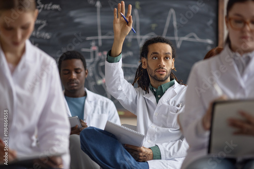 Obraz na plátne Multiethnic young adult male medical student raising hand during classroom lesson, participating in discussion with diverse classmates