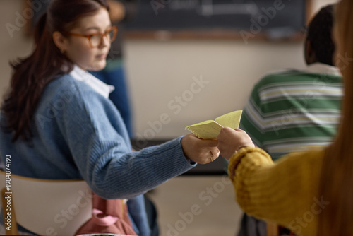 фотографія Teenage girl passing handwritten note to classmate during lesson, suggesting classroom distraction and peer communication