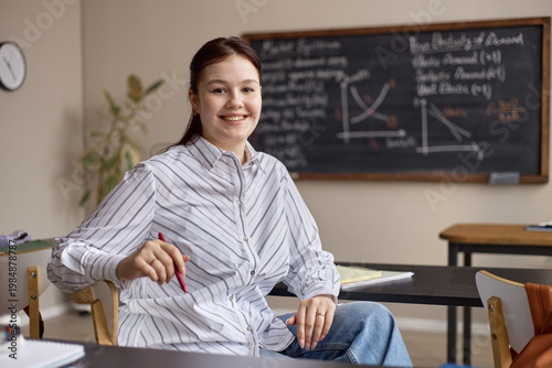 Φωτογραφία Portrait of Caucasian young adult woman sitting in classroom, holding pen, smiling at camera with math board behind