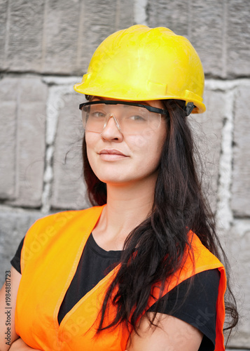 Young woman in yellow hard hat helmet, protective goggles and orange reflective vest, standing next to construction site bare wall