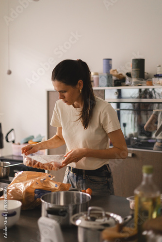 Young woman in a modern kitchen opening a plastic vacuum package of raw chicken meat. Female model preparing poultry for cooking healthy dinner at home, authentic lifestyle scene.