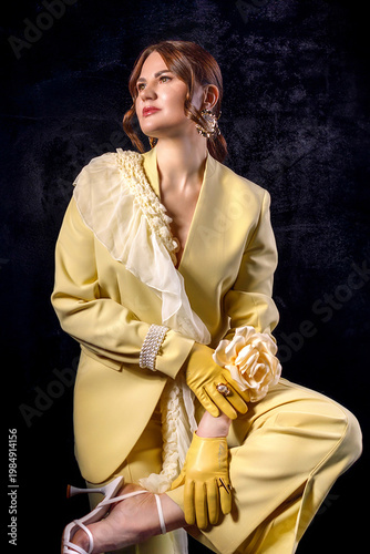 High fashion photography: a beautiful, elegant woman in a yellow pantsuit posing against a dark background. Slim figure. Hair cut and styled. Studio shot. The model is seated on a chair.