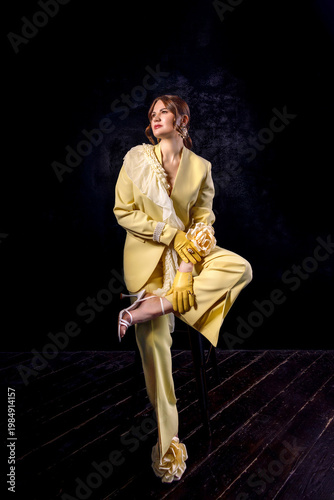 High fashion photography: a beautiful, elegant woman in a yellow pantsuit posing against a dark background. Slim figure. Hair cut and styled. Studio shot. The model is seated on a chair.