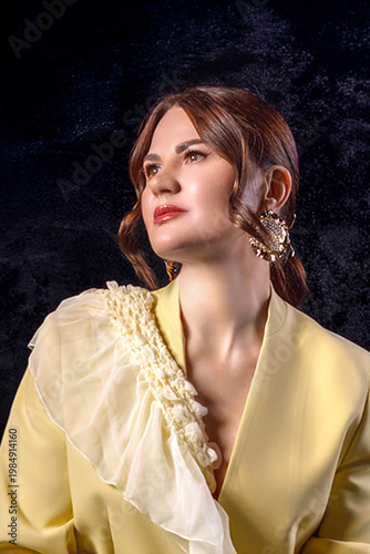 High fashion photography: a beautiful, elegant woman in a yellow pantsuit posing against a dark background. Slim figure. Hair cut and styled. Studio shot. The model is seated on a chair.