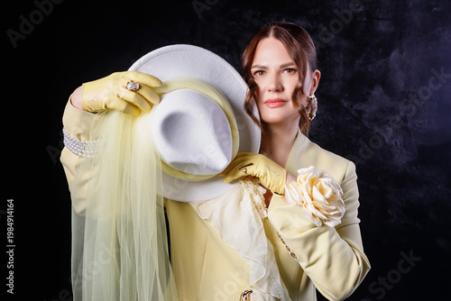 High fashion photography: a beautiful, elegant woman in a yellow pantsuit posing against a dark background. Slim figure. Hair cut and styled. Studio shot. The model is seated on a chair.