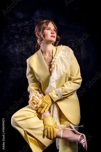 High fashion photography: a beautiful, elegant woman in a yellow pantsuit posing against a dark background. Slim figure. Hair cut and styled. Studio shot. The model is seated on a chair.