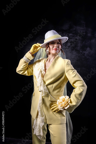 High fashion photography: a beautiful, elegant woman in a yellow pantsuit posing against a dark background. Slim figure. Hair cut and styled. Studio shot. The model is seated on a chair.
