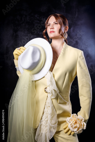 High fashion photography: a beautiful, elegant woman in a yellow pantsuit posing against a dark background. Slim figure. Hair cut and styled. Studio shot. The model is seated on a chair.