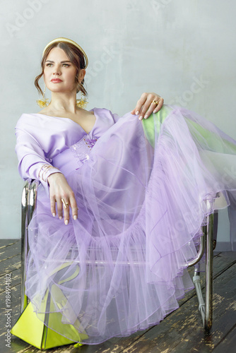A beautiful, elegant woman in a lilac dress, bright green neon boots, and a handbag posing against a light background. Slender figure. Hair cut and styled. Studio shot. The model is seated on a chair.