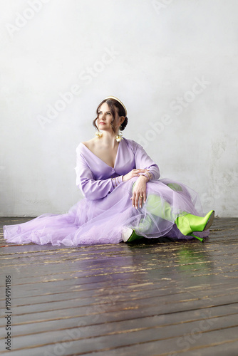 A beautiful, elegant woman in a lilac dress, bright green neon boots, and a handbag posing against a light background. Slender figure. Hair cut and styled. Studio shot. The model is seated on a chair.