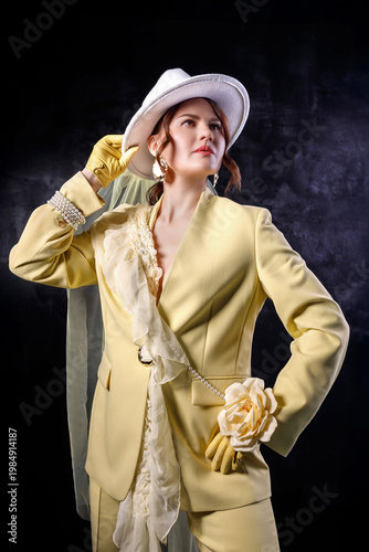 High fashion photography: a beautiful, elegant woman in a yellow pantsuit posing against a dark background. Slim figure. Hair cut and styled. Studio shot. The model is seated on a chair.