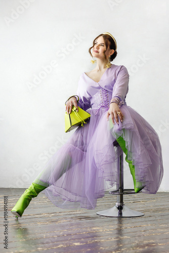 A beautiful, elegant woman in a lilac dress, bright green neon boots, and a handbag posing against a light background. Slender figure. Hair cut and styled. Studio shot. The model is seated on a chair.
