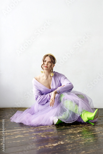 A beautiful, elegant woman in a lilac dress, bright green neon boots, and a handbag posing against a light background. Slender figure. Hair cut and styled. Studio shot. The model is seated on a chair.
