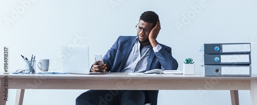 Tableau sur toile A young man is sitting at his desk in an office looking worried while holding his smartphone