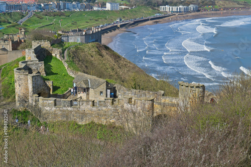 Ancient castle ruins stand on a green cliff overlooking a sandy beach with waves and a coastal town in Scarborough Castle - Yorkshire - Great Britain