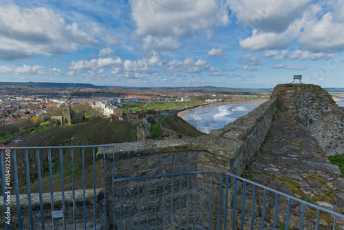 A wide scenic view from stone castle walls and railings, overlooking a town, a sandy beach, and the ocean under a blue, cloudy sky in Scarborough Castle - Yorkshire - Great Britain
