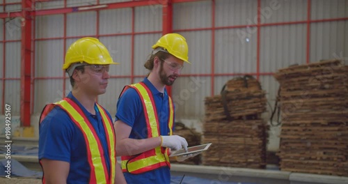 Two factory workers conduct a site inspection using a digital tablet to manage logistics and safety compliance inside a large industrial warehouse.