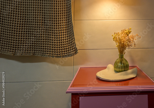 Dried flowers in green vase on white pottery stand on pink nightstand with checkered lampshade in soft indoor lighting. Cozy indoor furniture.