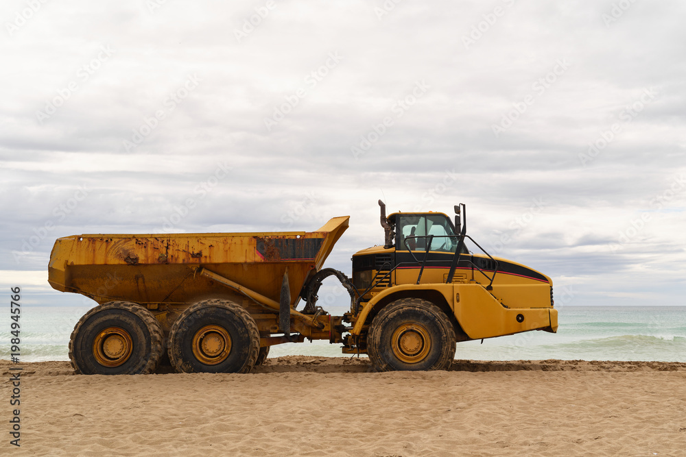 Obraz premium Large yellow dump truck standing on sandy beach near the sea under cloudy sky. Heavy machinery used for coastal work.
