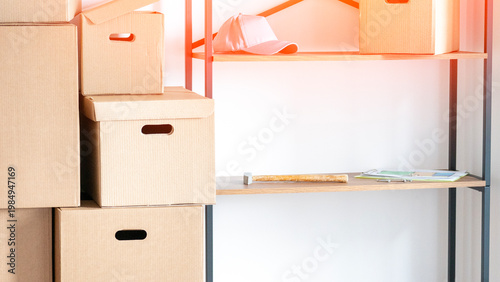 Cardboard boxes stack on metal shelves. White cap rests on an upper shelf. Ruler and paper lie on a lower shelf. Clean storage setup for office spaces. Perfect for organization or moving concepts