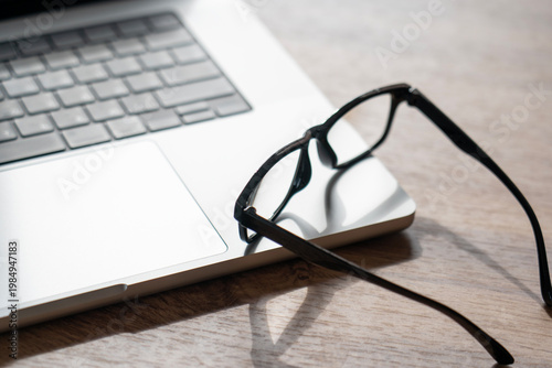 Silver laptop keyboard sits on a wooden table. Black eyeglasses lean against the laptop's trackpad. Natural light creates a serene workspace mood. This setup implies concentration during work or study