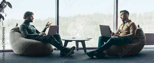 Fotografie Two men sit facing each other in an office space with large windows
