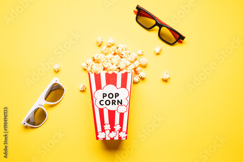 Popcorn with cheese taste in white and red cardboard bucket and 3d glasses on bright yellow background top view.