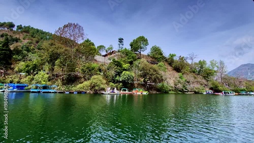 A serene lakeside scene with gently rippling green water, colorful boats lined along shore, and a lush hillside rising above, crowned by a small watchtower overlooking landscape.