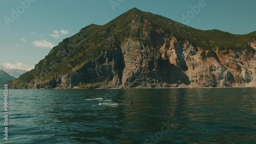 Pod of killer whales emerges from water in unison and releasing plumes of steam. View of steep mountain ranges overlooking water.