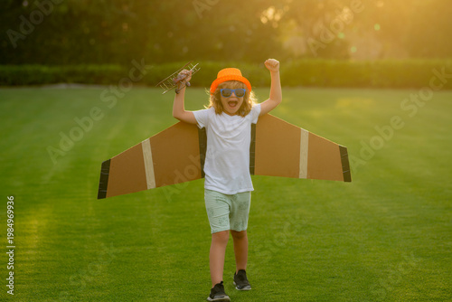 Kid pilot with paper wings or toy airplane having fun on meadow. Pilot aviator child with a toy airplane plays on summer nature. Child playing with plane wings. Concept of dreams and travels.